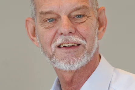 Allan Holm Pedersen, en mand med kort gråt hår og trimmet skæg, er iført en lyseblå skjorte med krave og smiler foran en almindelig lys baggrund.