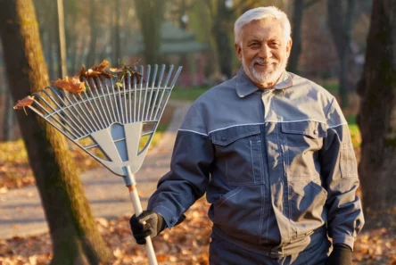 En ældre mand i grå jakke og handsker smiler, mens han holder en rive med blade i en park om efteråret og omfavner sin havn i november. Nedfaldne blade dækker jorden, og man kan se træer i baggrunden.