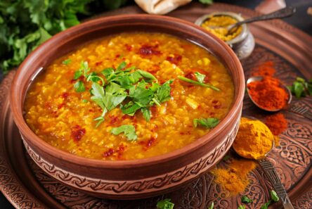 En skål med gul linsedal, en klassisk indisk vegetarret, pyntet med frisk koriander og omgivet af krydderier på en dekorativ tallerken. Denne varme og mættende ret viser nem vegetarret og krydret indisk mad med et drys af røde krydderier på toppen.