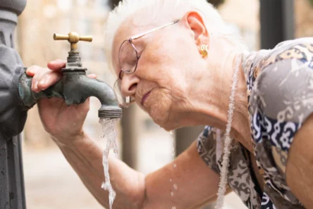 En ældre kvinde drikker vand fra en offentlig udendørs vandhane i København, hvor hun læner sig frem for at nå vandstrålen med munden – et enkelt øjeblik, der afspejler traditionen med gratis drikkevand i København til alle, der har brug for forfriskning.