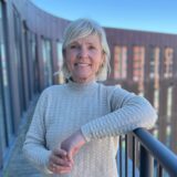 En smilende ældre kvinde, Birgitte Rømer, med kort blondt hår og en lys sweater, står udendørs på en balkon med moderne bygninger og en blå himmel i baggrunden.