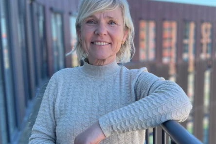 En smilende ældre kvinde, Birgitte Rømer, med kort blondt hår og en lys sweater, står udendørs på en balkon med moderne bygninger og en blå himmel i baggrunden.