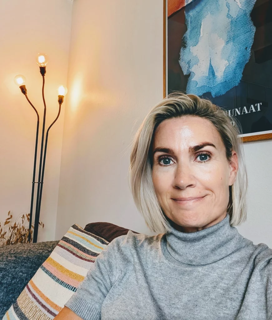En ældre kvinde med kort blondt hår og en grå rullekravesweater sidder på en sofa med stribede puder. I baggrunden ses en moderne gulvlampe med tre pærer og abstrakt kunst, hvilket skaber en hyggelig ramme for et foredrag om sorgbearbejdelse.