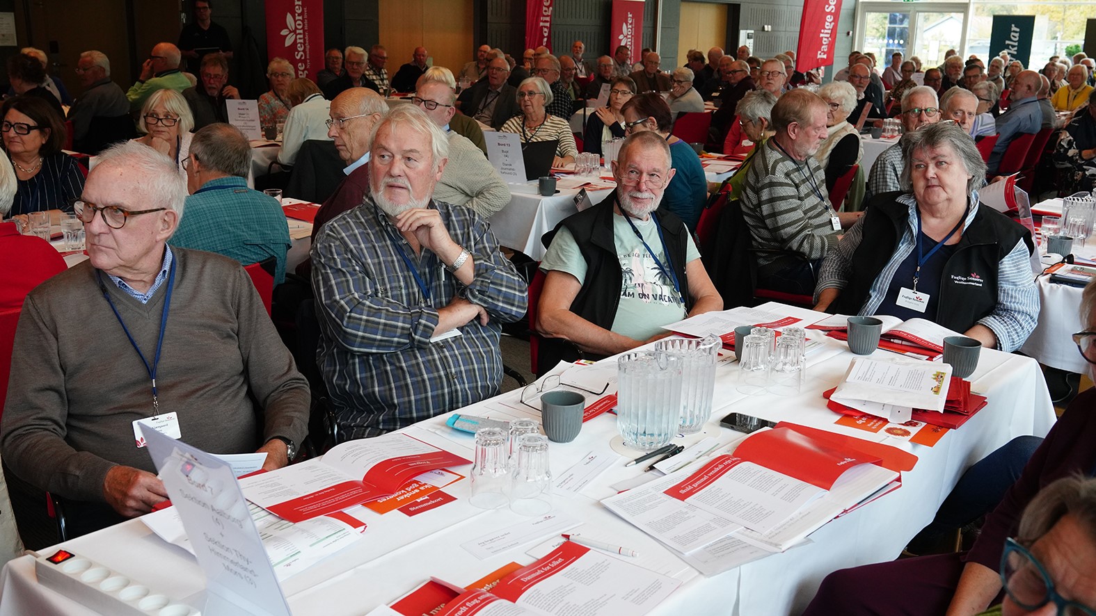 En stor gruppe seniorer fra Faglige Seniorer sidder ved runde borde dækket af papirer, pjecer og vandkander og deltager i en veloplyst konference. Mange af deltagerne bærer navneskilte og lytter opmærksomt, mens der diskuteres nyhedsbreve.