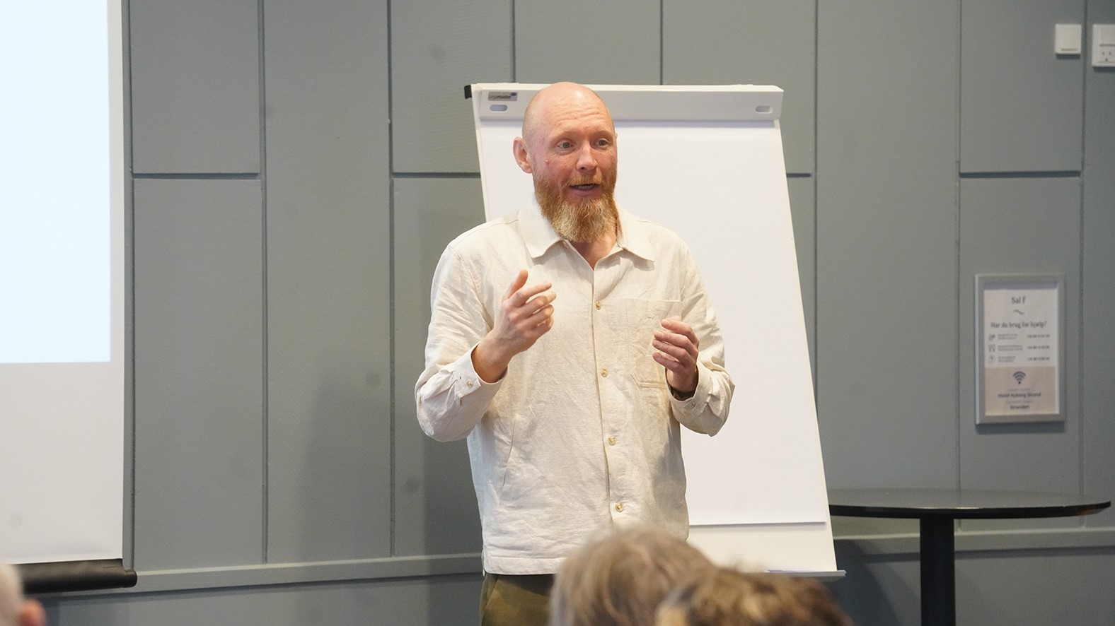 En mand med skaldet hoved og rødligt skæg, sandsynligvis en forsker, iført en lys skjorte, taler om ældrediskrimination foran et publikum, der står ved en flipover i et moderne konferencelokale.