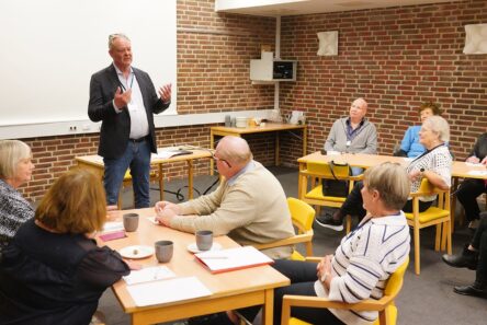 En mand står og taler til en lille gruppe seniorer, der sidder omkring borde i et klasseværelse med murstensvægge og et projektorlærred i baggrunden. Gruppen lytter opmærksomt i trygge rammer.