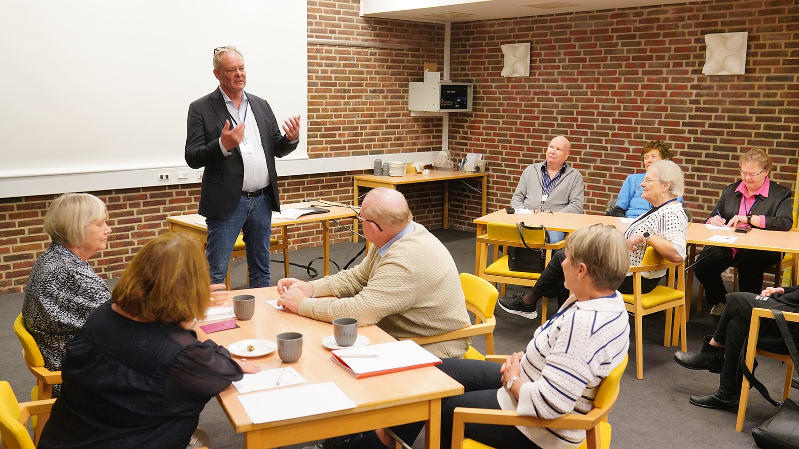 En mand står og taler til en lille gruppe seniorer, der sidder omkring borde i et klasseværelse med murstensvægge og et projektorlærred i baggrunden. Gruppen lytter opmærksomt i trygge rammer.