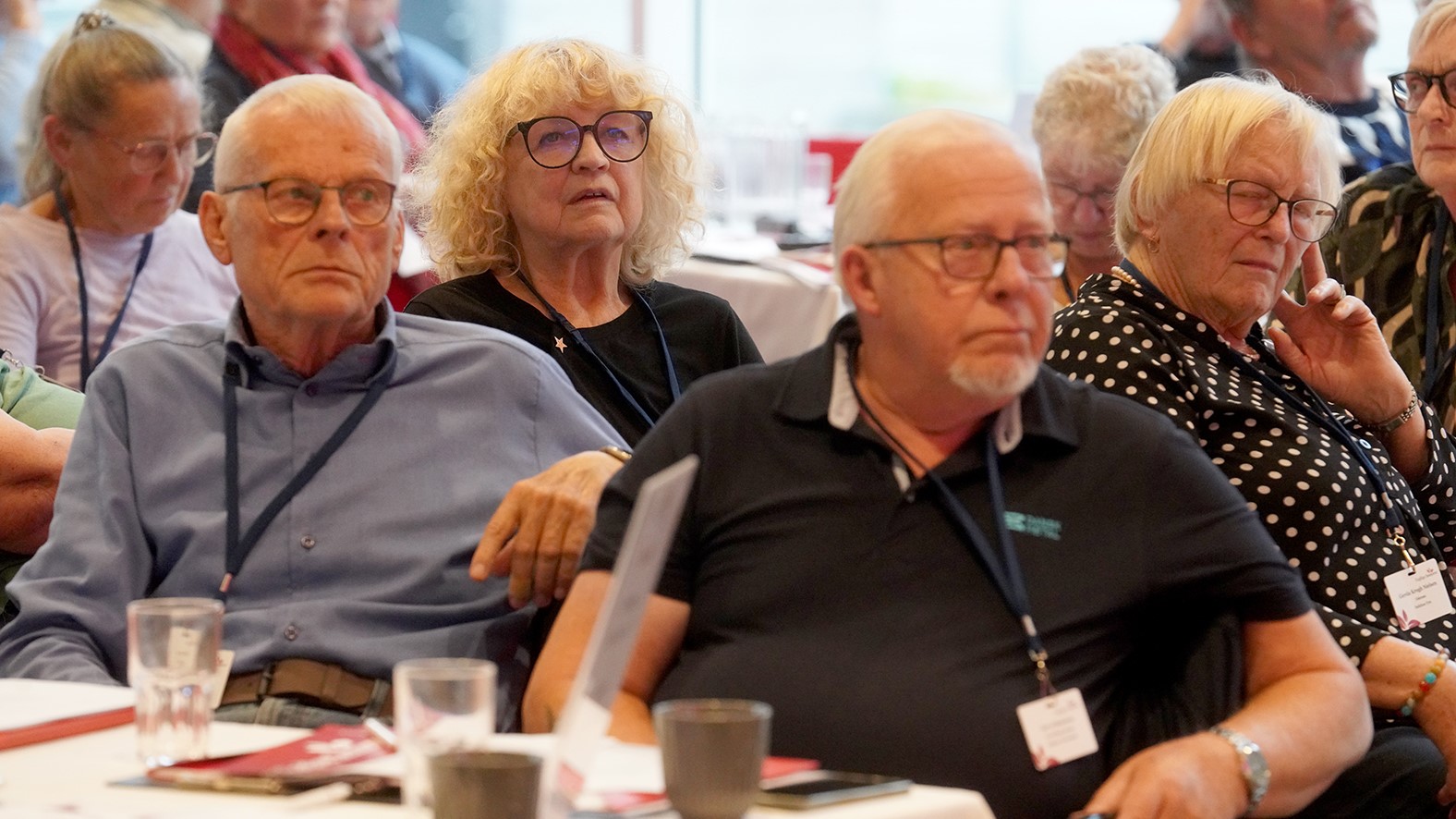 En gruppe ældre voksne sidder tæt sammen ved bordene og lytter opmærksomt til en debat under et indendørs arrangement. Nogle bærer briller og navneskilte, mens notesbøger og kopper er synlige på bordene, mens de følger med i beretningen.