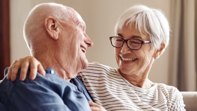 Et ældre par sidder tæt sammen i en sofa og smiler kærligt til hinanden, som om de deler en hemmelighed om aftalen. Kvinden har briller og en stribet skjorte på, mens manden har en blå skjorte på.