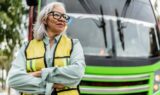 En selvsikker ældre kvinde med gråt hår og briller, iført en gul sikkerhedsvest, står med korslagte arme foran en lysegrøn bus - et stærkt eksempel på seniorer der arbejder.