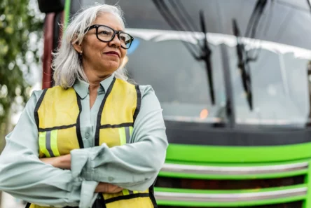 En selvsikker ældre kvinde med gråt hår og briller, iført en gul sikkerhedsvest, står med korslagte arme foran en lysegrøn bus - et stærkt eksempel på seniorer der arbejder.