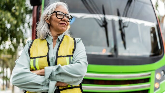 En selvsikker ældre kvinde med gråt hår og briller, iført en gul sikkerhedsvest, står med korslagte arme foran en lysegrøn bus - et stærkt eksempel på seniorer der arbejder.