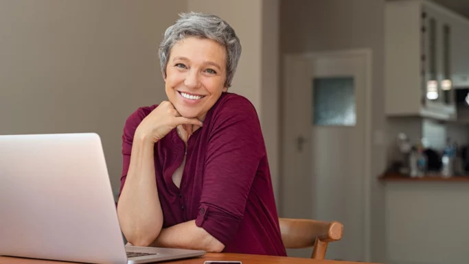 En smilende ældre kvinde med kort gråt hår sidder ved et bord og bruger en bærbar computer. Hun har en rødbrun skjorte på og hviler hagen på sin hånd, mens hun kigger ind i kameraet. Baggrunden viser et køkkenområde, hvilket understreger, hvordan seniorer omfavner teknologi.