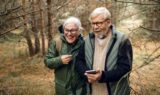 Et ældre par iført briller og jakker smiler og griner, mens de går sammen gennem et skovområde og holder en smartphone. Tørre blade og fyrretræer omgiver dem og fremhæver betydningen af fælles gåture for at skabe glæde og samhørighed.