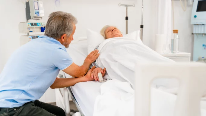 En mand sidder ved siden af en hospitalsseng og holder forsigtigt hånden på en ældre patient, der ligger under hvide lagner, omgivet af medicinsk udstyr i et lyst rum - og afspejler øjeblikke, der er formet af Covid-19 og bekymringer om dødelighed.