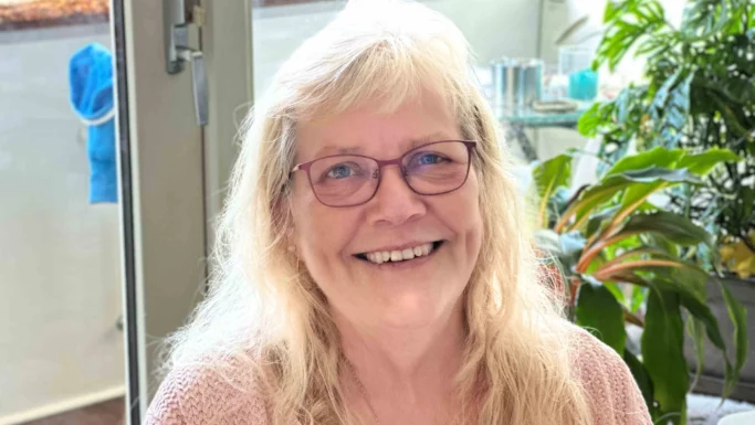 En smilende ældre kvinde med langt blondt hår og briller sidder indendørs nær grønne planter og et vindue iført en lyserød sweater. Hendes varme udtryk afspejler erfaring og et ønske om at hjælpe andre. Klart naturligt lys fylder rummet.