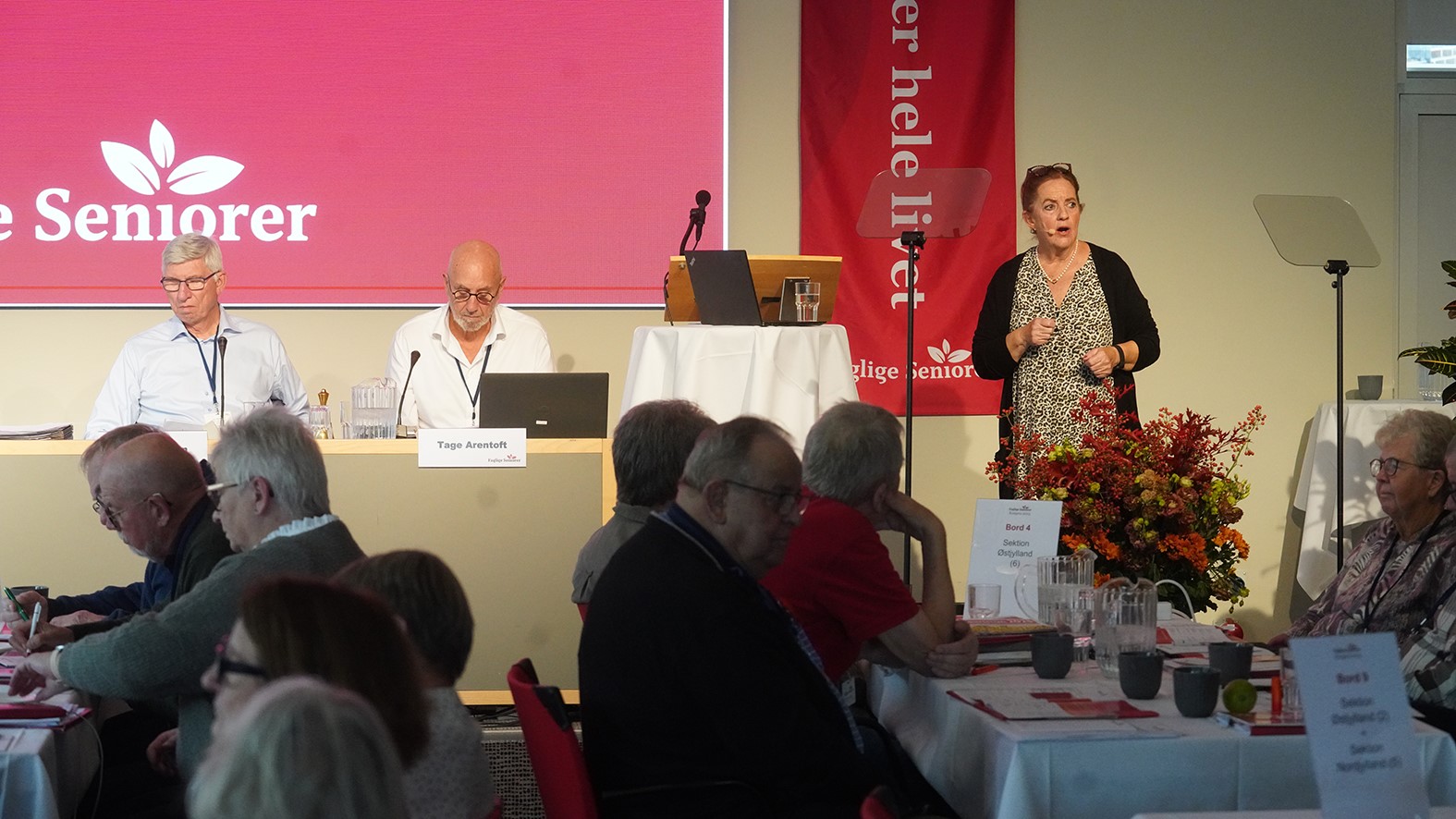 En kvinde, muligvis Mette Kindberg, taler til et siddende publikum ved et seniorarrangement. To mænd med bærbare computere sidder på scenen. Et rødt banner og et blomsterarrangement tilføjer farve, mens folk i fællesskabet lytter og tager noter i forgrunden.