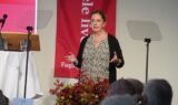 En kvinde taler foran et publikum, stående mellem to gennemsigtige podieskærme. Hun repræsenterer Faglige Seniorer og er iført en sort cardigan over en mønstret bluse med et rødt banner og blomster i baggrunden.