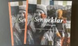 To eksemplarer af SeniorKlarhåndbogen med foto af en smilende ældre kvinde udendørs, iført jakke og hue, og en ældre mand i baggrunden. Det farverige omslag giver overblik og inspiration til et aktivt seniorliv.