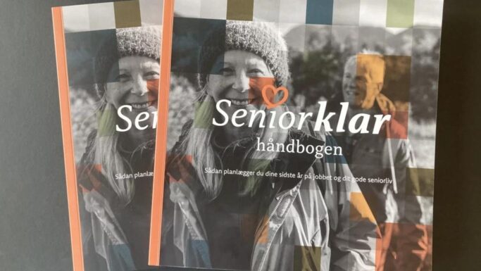 To eksemplarer af SeniorKlarhåndbogen med foto af en smilende ældre kvinde udendørs, iført jakke og hue, og en ældre mand i baggrunden. Det farverige omslag giver overblik og inspiration til et aktivt seniorliv.