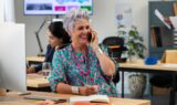 En smilende ældre kvinde med kort, gråt hår taler i telefon ved sit skrivebord på et moderne kontor. Hun er iført en farverig grøn og pink kjole og briller, mens en kollega arbejder i baggrunden.