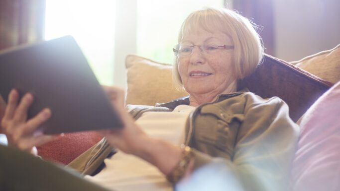 En ældre kvinde med briller slapper af i en sofa og smiler, mens hun bruger en tablet. Sollyset strømmer ind fra et vindue bag hende og skaber en varm og behagelig atmosfære.