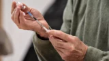 En person i en grøn sweater holder et hætteglas og forbereder en sprøjte, hvor han trækker væske ud af hætteglasset, muligvis til vaccination, mens han tjekker vaccineringsfakta for at sikre korrekt procedure og undgå fejl.