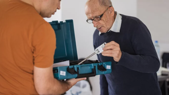 En ældre mand med briller undersøger et stykke værktøj fra en blågrøn værktøjskasse, som holdes af en anden person i orange skjorte. Det ser ud til, at de praktiserer deleøkonomi og diskuterer, hvordan man kan dele ting med naboerne sammen indendørs.