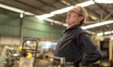 En ældre mand i arbejdstøj står på en fabrik, krummer ryggen og holder sig i lænden i smerte - et tydeligt tegn på det hårde arbejdsmiljø, som mange på seniorarbejdsmarkedet står over for. I baggrunden ses industrielt udstyr og slørede arbejdere.