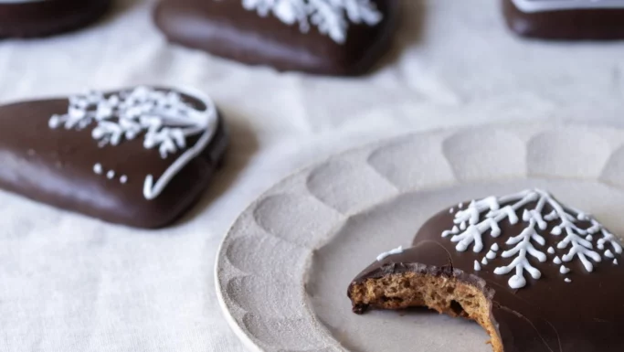 En hjerteformet honninghjerterkage med hvid glasur og en bid taget ud ligger på en tallerken. I baggrunden ligger lignende gode honninghjerter pyntet med hvid glasur på en lys dug.
