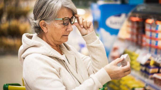 En ældre kvinde med briller holder et fødevareprodukt og læser på etiketten i en købmandsforretning, mens hun ser ud til at undersøge emballagen nøje.