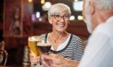 En ældre kvinde med kort gråt hår og briller smiler, mens hun holder et glas øl og nyder alkohol i en hyggelig, varmt oplyst bar, mens hun sidder over for en ældre mand med en mørk drink.