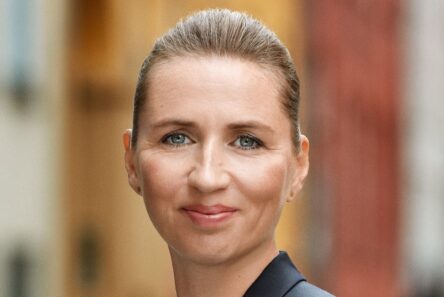 En kvinde med lys hud og blå øjne, der smiler blidt, har sit lysebrune hår trukket tilbage. Hun er iført en mørk blazer og står udendørs i København med slørede farverige bygninger i baggrunden.