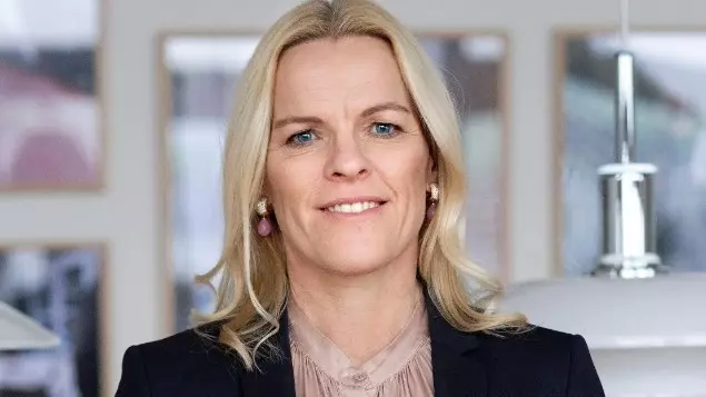 En kvinde med langt blondt hår, iført mørk blazer og lys bluse, står indendørs og smiler til kameraet. Indrammede kunstværker og moderne hængelamper antyder, at hun måske er på besøg på et plejehjem for at diskutere værdig pleje for beboerne.