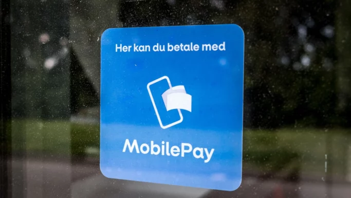 Et blåt klistermærke på glas viser et smartphone- og kort-ikon med teksten Her kan du betale med MobilePay, som viser, at disse butikker tilbyder kontaktløs betaling med MobilePay.