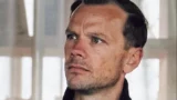 En mand med kort brunt hår og blå øjne kigger let opad med et alvorligt udtryk, mens han står indendørs ved et vindue med sløret lys, der reflekterer over justitsministerens arbejde mod svindel mod ældre og behov for hjælp.