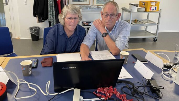To ældre voksne sidder tæt sammen ved et bord og kigger intenst på en Lenovo-laptop under et AI-kursus. På bordet ligger papirer, snacks, telefoner og navnekort, og i baggrunden står en kaffevogn med forsyninger.