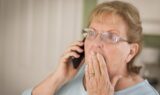 En ældre kvinde med briller holder en telefon op til øret med den ene hånd og holder sig for munden med den anden, mens hun ser chokeret ud - muligvis reagerer hun på nyheder om netbanksvindel. Hun befinder sig indendørs med en sløret baggrund.