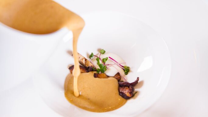 En cremet brun champignonsovs hældes fra en hvid kande op på et fad med grillede svampe, mikrogrønt og en klat hvid puré - alt sammen kunstfærdigt arrangeret på en hvid tallerken med metalaccent mod en sprød hvid baggrund.