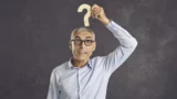 En ældre mand med briller og lyseblå skjorte holder et spørgsmålstegn af træ over hovedet og kigger op med et nysgerrigt udtryk - og fanger dermed ånden i spørgsmål og svar om folkepension og de ændringer, der kommer i folkepension 2026.