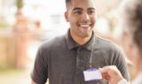 En smilende mand i en mørk polotrøje holder et blankt ID-badge frem til en ældre person, som rækker ud efter det. Det ser ud til, at de interagerer udendørs i nærheden af huse i en af byerne.