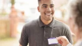 En smilende mand i en mørk polotrøje holder et blankt ID-badge frem til en ældre person, som rækker ud efter det. Det ser ud til, at de interagerer udendørs i nærheden af huse i en af byerne.