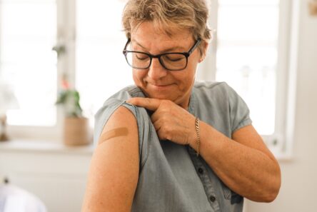 En ældre kvinde med briller smiler, mens hun peger på en bandage på sin overarm, som viser, at hun for nylig er blevet vaccineret mod influenza. Hun er indendørs og klædt i en kortærmet skjorte.