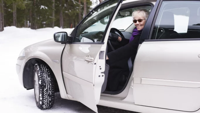 En ældre kvinde iført solbriller og et lilla tørklæde sidder på førersædet af bilen med døren åben. Den er parkeret på en snedækket vej og er vigtig for at sikre, at dæk er klar til vinteren med træer synlige i baggrunden.