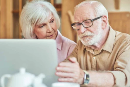 En ældre mand og kvinde sidder sammen ved et bord og kigger intenst på en laptop-skærm - måske for at gennemgå deres formue eller pensionstillæg. Manden gestikulerer mod skærmen, mens kvinden følger nøje med. En tekande og kopper står på bordet i forgrunden.