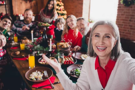 En smilende ældre kvinde tager en selfie med sin familie omkring et festligt julebord, hvor de nyder billig julemiddag med stearinlys og pynt. Et juletræ lyser op i baggrunden, mens de deler ekstra penge juletips.