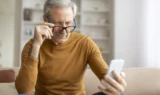 En ældre mand i en brun sweater sidder i en sofa og kigger intenst på sin mobil, mens han justerer sine briller. Den svagt oplyste, moderne stue fremhæver hans fokus, måske på at justere tekststørrelse eller kontrast for at gøre det lettere at læse.