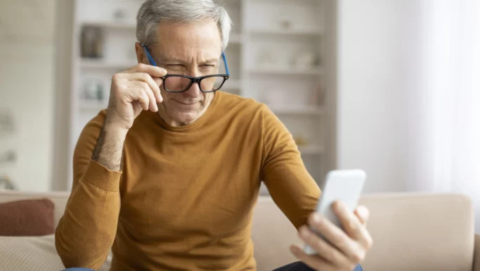 En ældre mand i en brun sweater sidder i en sofa og kigger intenst på sin mobil, mens han justerer sine briller. Den svagt oplyste, moderne stue fremhæver hans fokus, måske på at justere tekststørrelse eller kontrast for at gøre det lettere at læse.