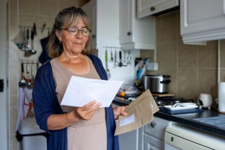 En ældre kvinde med briller står i et køkken og holder og læser et brev med en konvolut i den anden hånd. Hun er iført en beige kjole og blå cardigan og ser ud til at være fokuseret på brevets indhold.
