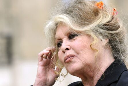 En ældre kvinde med bølget, lyst hår prydet med små orangefarvede blomster rører ved sit øre. Hun minder om Brigitte Bardot, er iført en mørk top og har et eftertænksomt udtryk på en sløret, neutral baggrund.